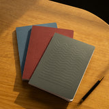 The Layflat Notebook Bundle (Series 2 - Set of 3) sits stacked on a wooden surface beside a black and gold pen, with sunlight casting soft shadows across the table.