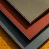 A stack of three Layflat Notebooks (Charcoal) with textured covers and Smyth sewn binding rests on wood, their visible spines highlighted by sunlight casting soft shadows.
