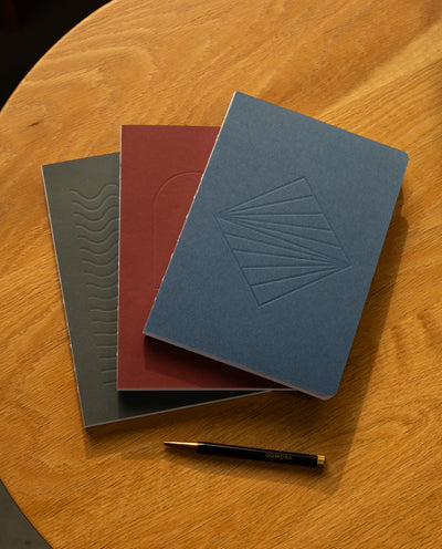 The Layflat Notebook Bundle (Series 2 - Set of 3) features textured gray, maroon, and blue notebooks arranged in a fan shape on a wooden table, with a black and gold pen placed below them.