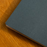 A close-up of a navy Layflat Notebook with dot grid pages and Smyth sewn binding, featuring the UGMONK debossed logo on the cover, resting on a wooden surface.