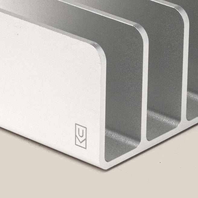 Close-up of a metallic object with three parallel, rectangular slots and beveled edges. The surface is smooth with a small, simple logo in the bottom left corner. The background is plain and light-colored.