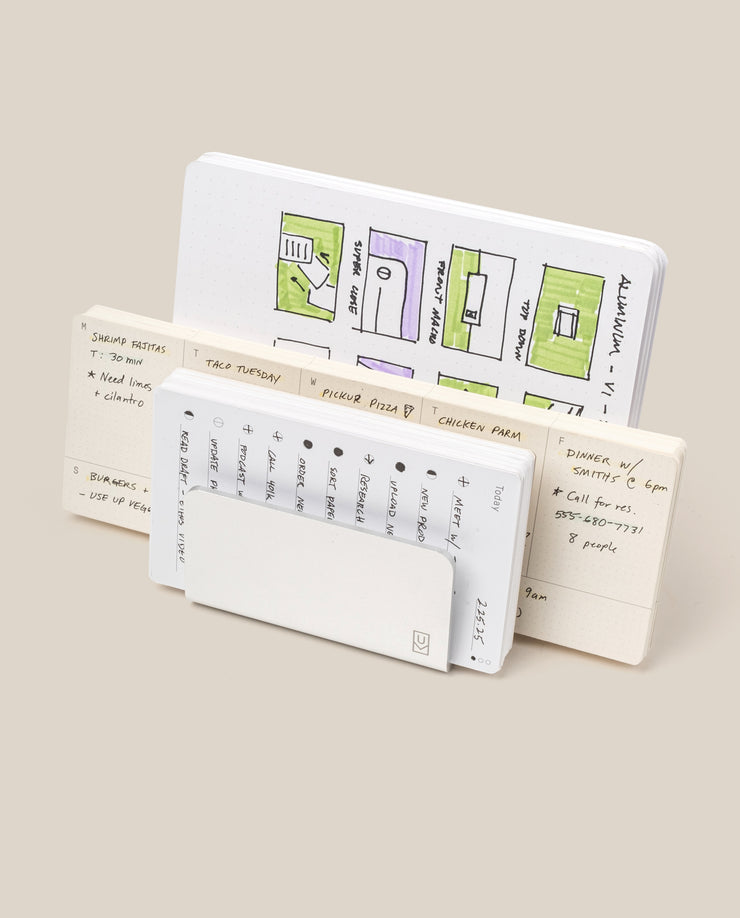 Three open notebooks stand upright. The back features colorful floor plans; the middle, handwritten notes and a meal plan. In front, a small calendar peeks from its cover next to the Analog Card Organizer (Silver Aluminum) for sleek card storage.