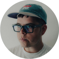 A person wearing a blue cap, black-rimmed glasses, and a white shirt looks slightly to the side. The background is plain and light-colored.