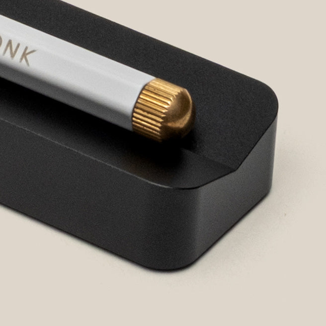 A close-up of a white pencil with a gold-colored eraser cap resting on a black rectangular holder against a beige background.