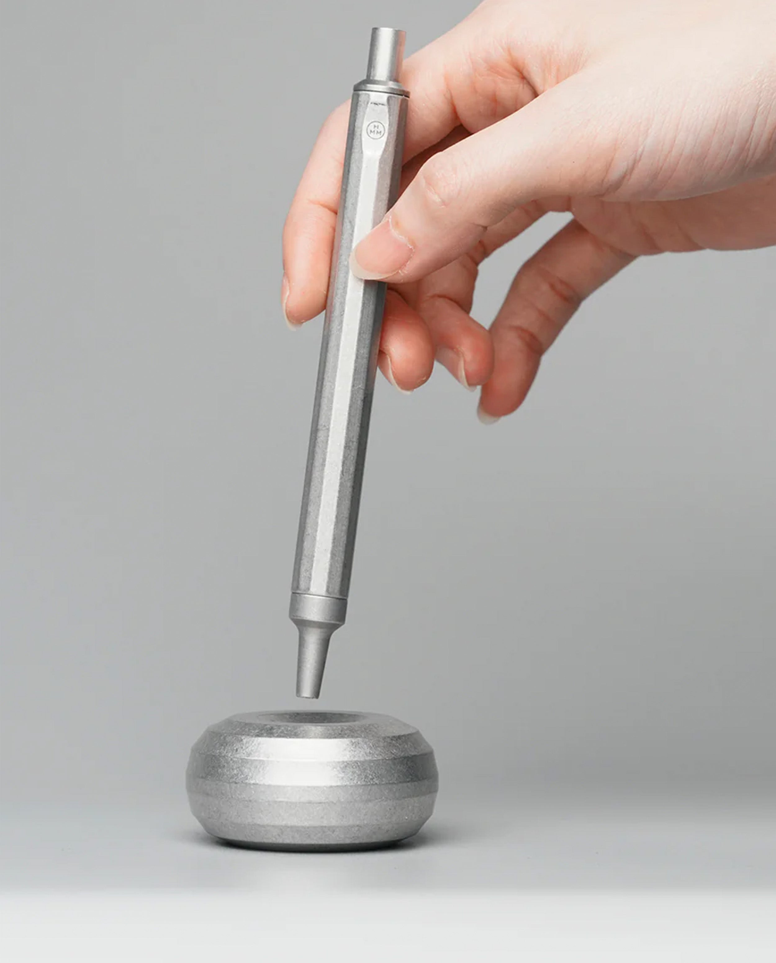 A hand holds the HMM Paperweight Pen Stand (Raw) vertically above its round, silver aluminum base on a gray background. The tip of the pen is near the center of the base, which serves as an elegant paperweight and a stylish desk accessory.