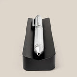 A pen from The Complete Analog Collection (Black Aluminum) with a ribbed grip lies horizontally on a black rectangular holder—an elegant desk accessory, shown tip-first against a plain beige background.