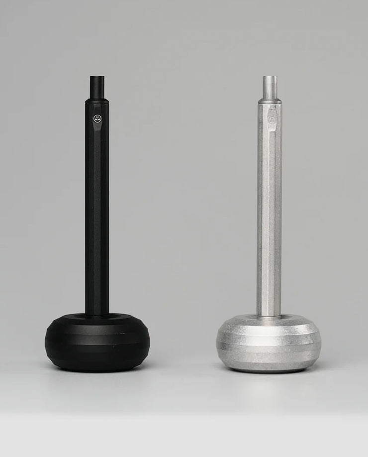 Two HMM Paperweight Pen Stands (Black), with hexagonal handles and round bases, are displayed side by side on a plain gray background, offering a quirky desk accessory that doubles as a pen stand or paperweight.