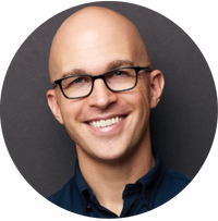 A smiling bald person wearing black glasses and a dark collared shirt, pictured against a plain dark background.