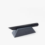 A black utility knife with a segmented blade rests on the Craighill Desk Knife Plinth (Slate), a geometric, wedge-shaped cast concrete holder, against a white background.