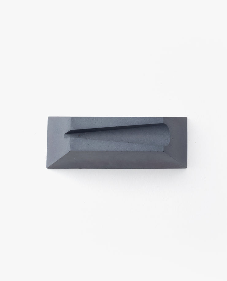 A rectangular, dark gray cast concrete Craighill Desk Knife Plinth (Slate) with a sloped, angular groove on top is shown as a sleek holder, set against a plain white background.