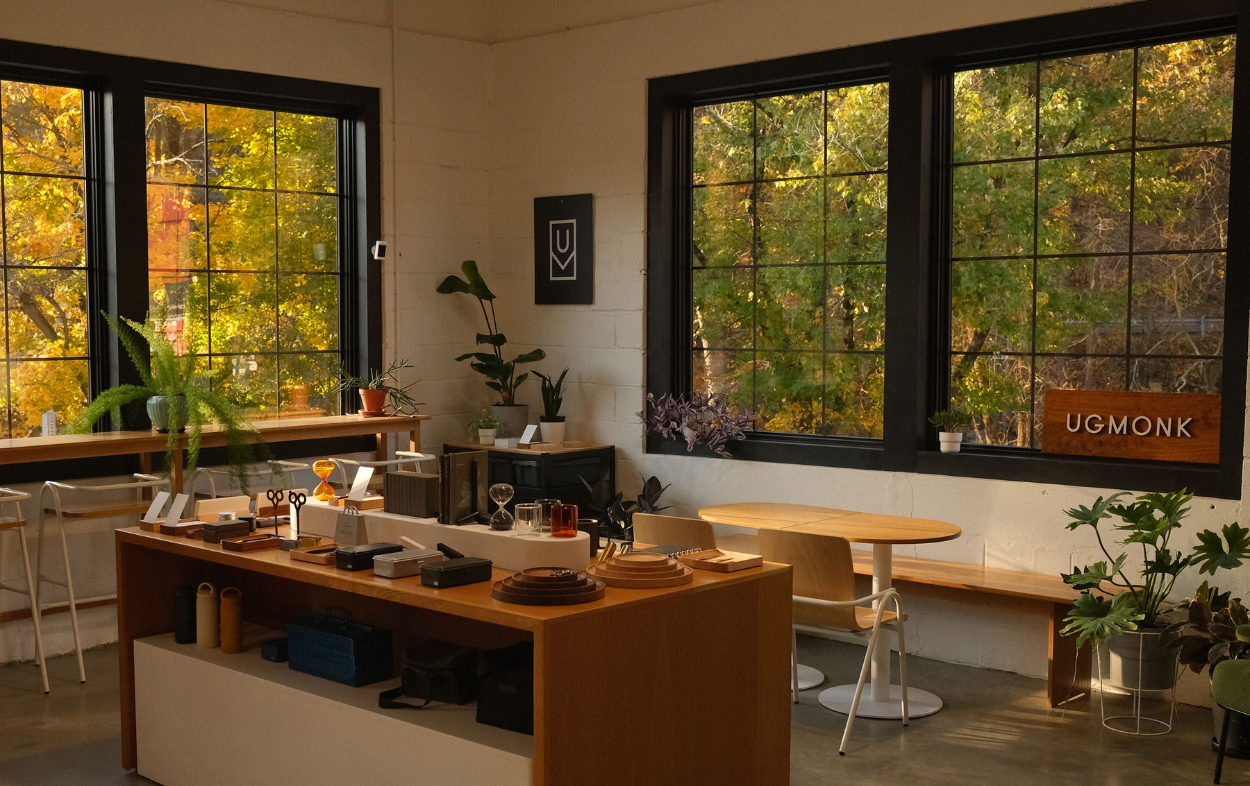 A cozy, sunlit shop with large windows shows trees with autumn foliage outside. Inside are shelves and tables displaying home goods, plants, and minimalist decor. A wooden UGMonk sign is visible by the window.