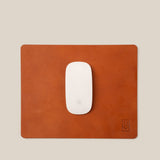 A white Apple Magic Mouse sits centered on the Ugmonk Leather Mousepad (Tan), handcrafted from full-grain leather with a subtle debossed logo in the lower right, all set against a neutral beige background.