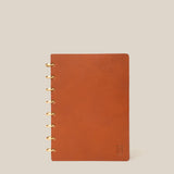 A Discbound Heirloom Journal in tan leather (6" x 8.5") with gold rings sits on a beige background. It features dot grid pages and a small embossed logo in the bottom right of the cover.