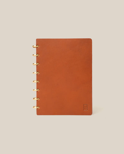 A Discbound Heirloom Journal in tan leather (6" x 8.5") with gold rings sits on a beige background. It features dot grid pages and a small embossed logo in the bottom right of the cover.