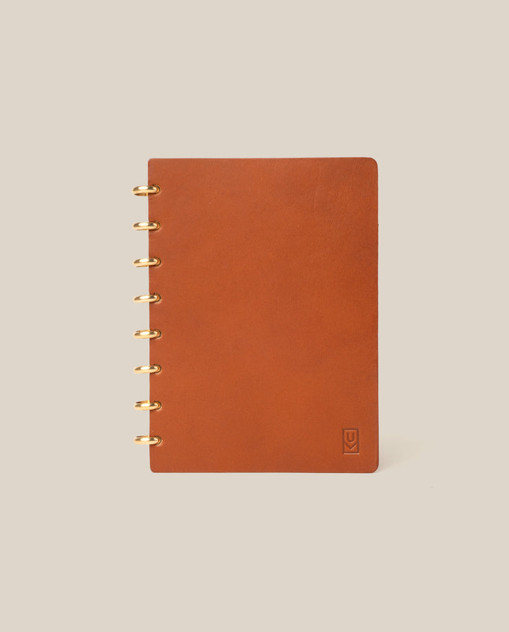 A Discbound Heirloom Journal in tan leather (6" x 8.5") with gold rings sits on a beige background. It features dot grid pages and a small embossed logo in the bottom right of the cover.