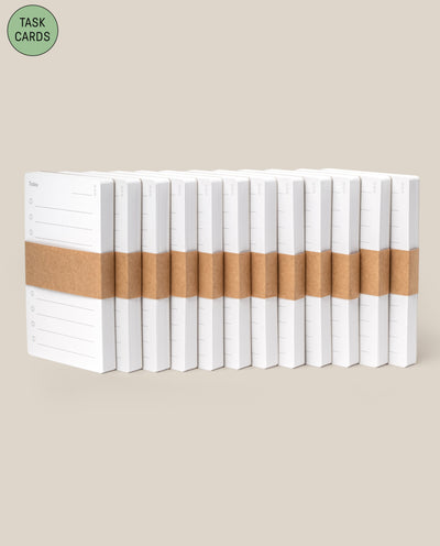 A pack of 12 Analog - Task Cards, each white notepad wrapped with a brown paper band, is arranged against a beige background. A green circle in the top left says TASK CARDS—ideal for any analog productivity system.