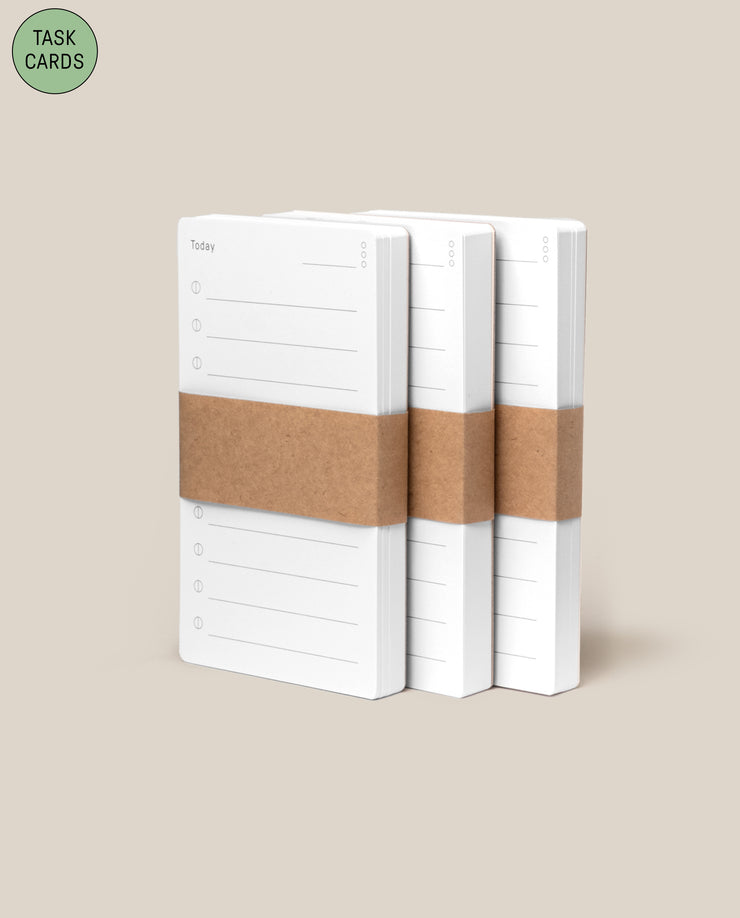 Three stacks of Analog - Task Cards (3-Pack), each wrapped in a brown paper band, sit side by side on a beige background. A green circle in the top left corner displays “TASK CARDS”—ideal for your analog productivity system.