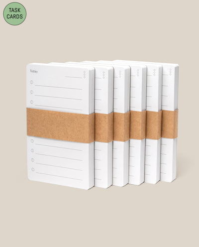 Six stacks of white Analog - Task Cards (6-Pack), an analog productivity tool, are bound with brown paper bands and arranged on a beige background. A green circle labeled "TASK CARDS" appears in the top left corner.