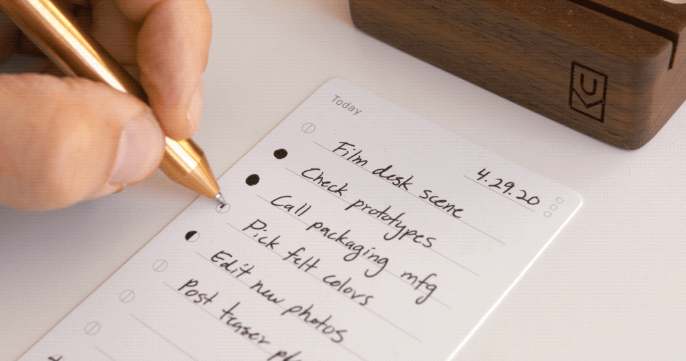 A hand holding a gold pen writes on a white to-do list. Tasks include “Film desk scene,” “Check prototypes,” and “Call packaging mfg.” The date “4.29.20” is written at the top of the list.