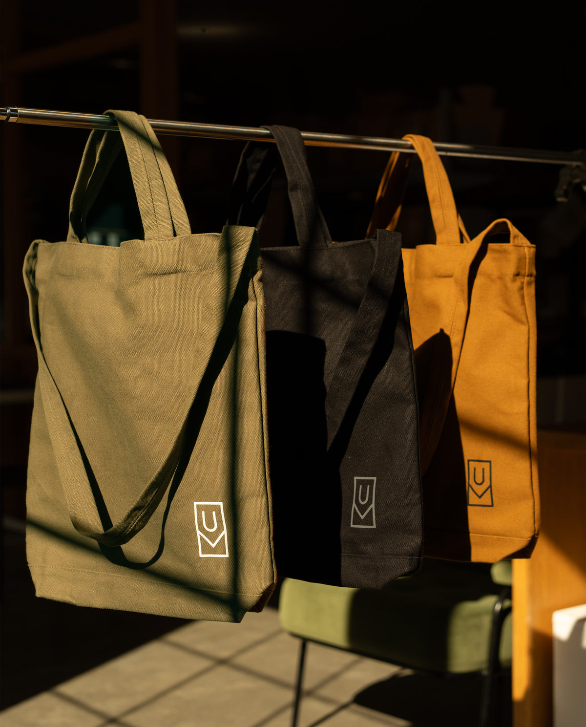 A Utility Tote Bundle - Set of 3 featuring three heavyweight cotton totes in olive, black, and mustard hanging from a metal rod, each with a small white logo on the front.