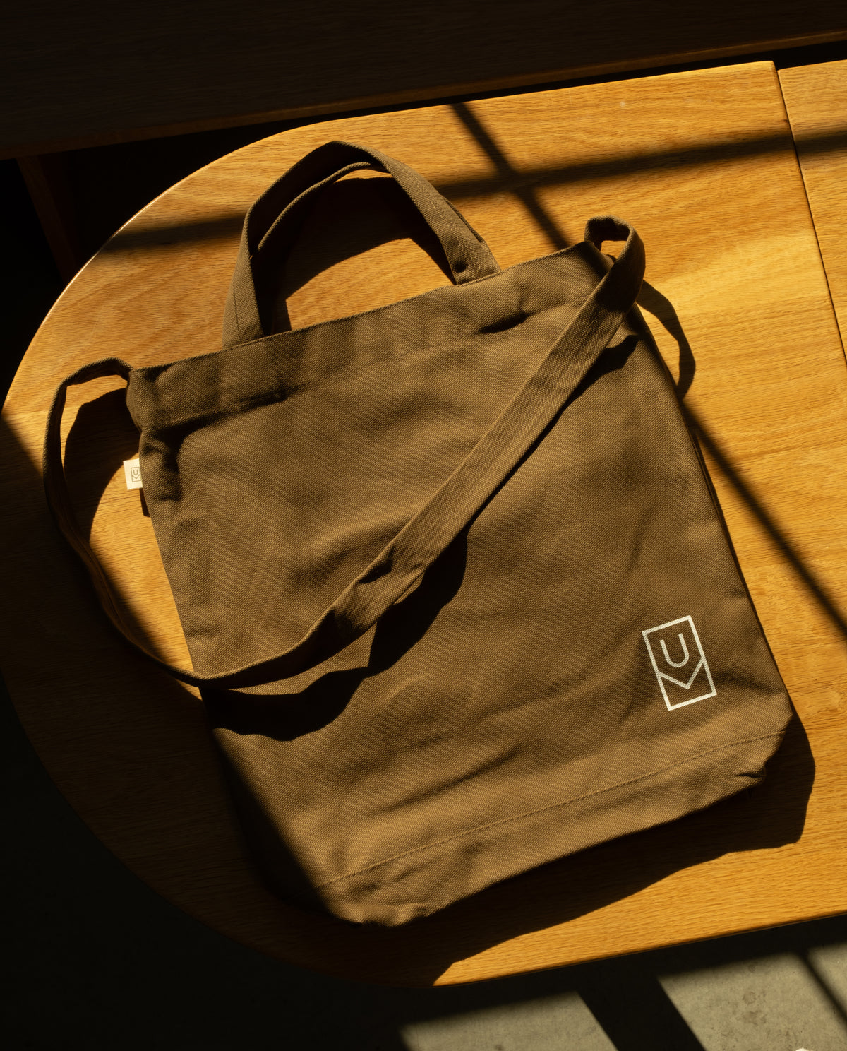 Sturdy olive green canvas Ugmonk Utility Tote Bundle (Set of 3) with a shoulder strap and a small geometric logo on the bottom corner, resting on a round wooden table.