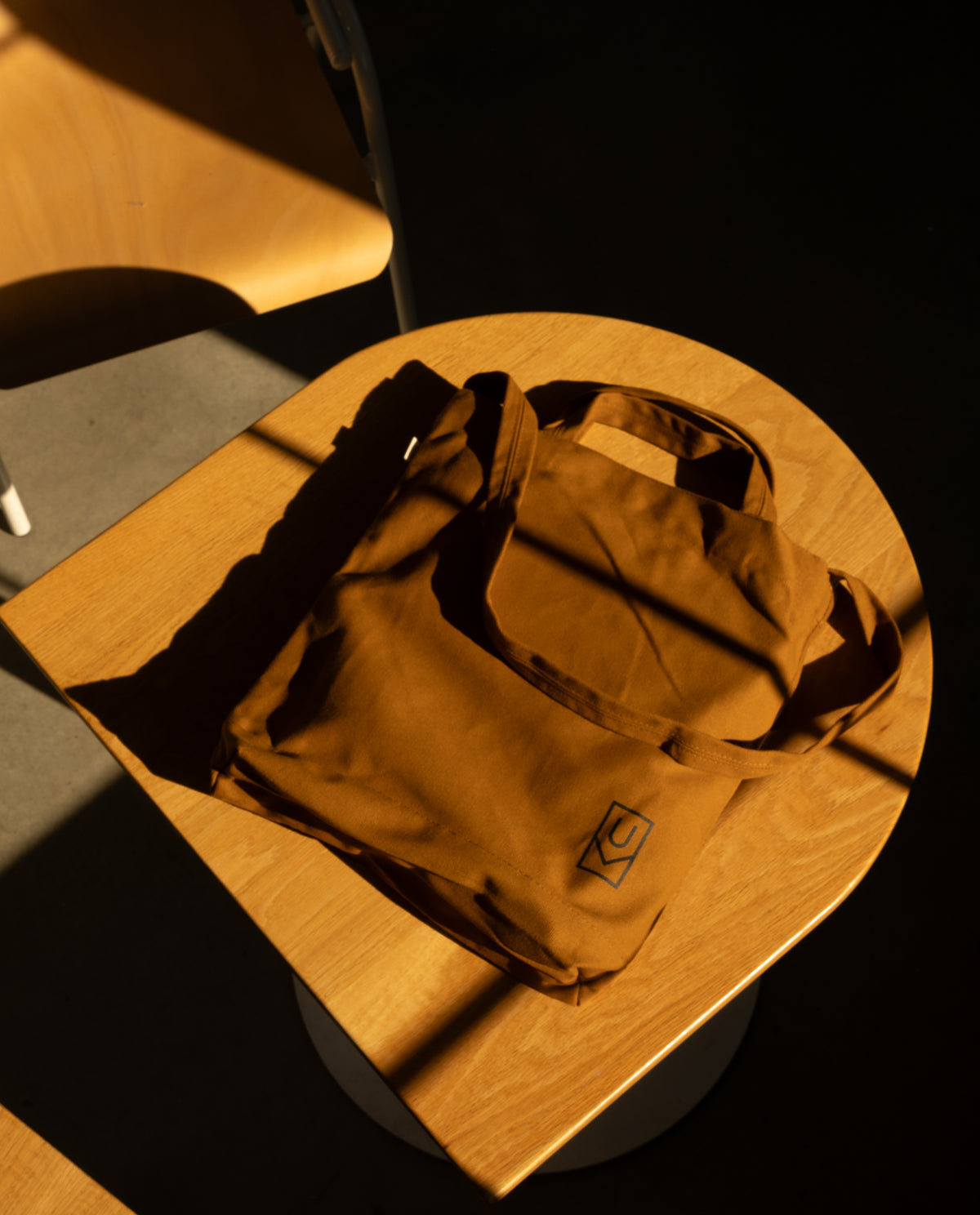 Ugmonk Utility Tote (Field Tan) heavyweight cotton utility tote bag with a small square logo, sitting on a wooden stool.