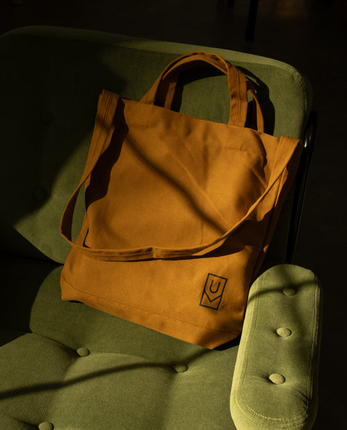 Ugmonk Utility Tote (Field Tan) made of sturdy tan canvas with a small black logo in the corner, resting on a green armchair.