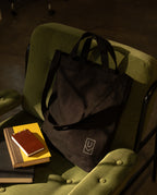 Black Ugmonk Utility Tote with a geometric logo resting on a green armchair next to a small notebook and a stack of books.