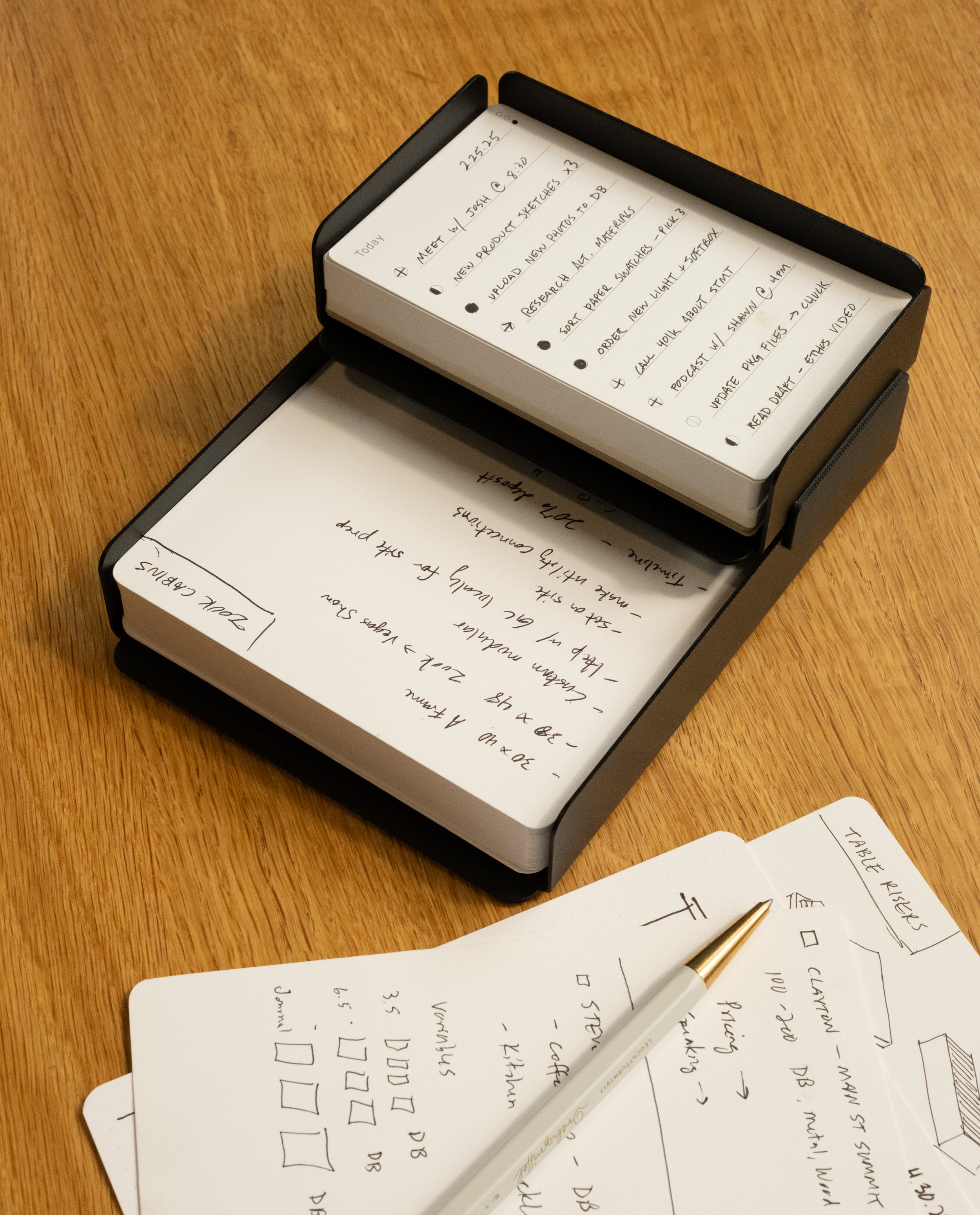 A stack of white Capture Cards with handwritten notes sits in a black holder on a wooden desk; nearby are more cards and a pen. The Capture Bundle (Small Tray + Large Tray - Black Steel) is ideal for organizing ideas with its stackable trays.