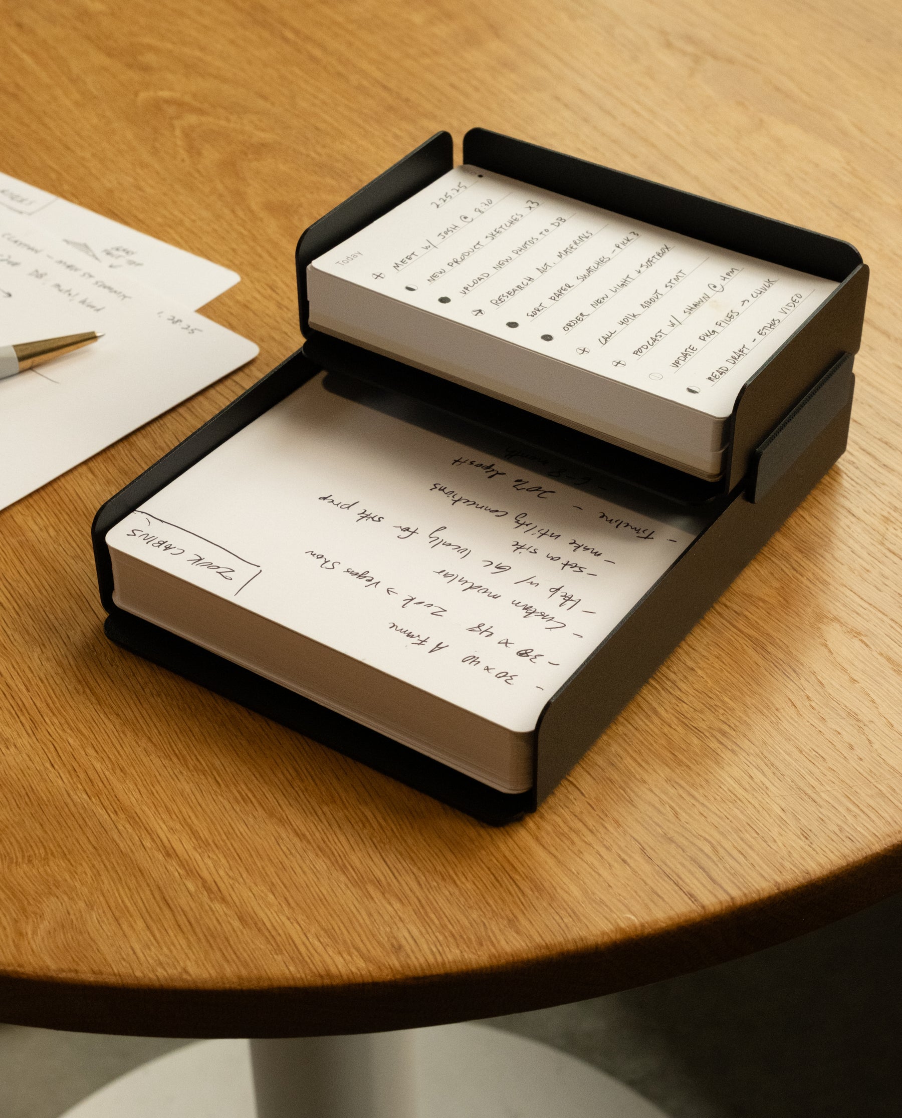 A black desk organizer holds two stacks of white notepaper filled with handwritten notes, placed on a wooden table next to a sheet of paper and a pen.