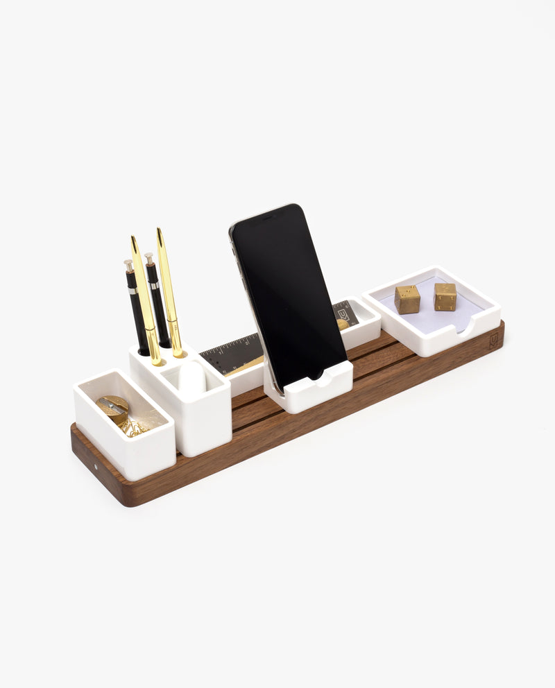 A wooden and white ceramic desk organizer holds pens, a smartphone, a ruler, binder clips, paper clips, and thumbtacks, all arranged neatly on a clean, white background.