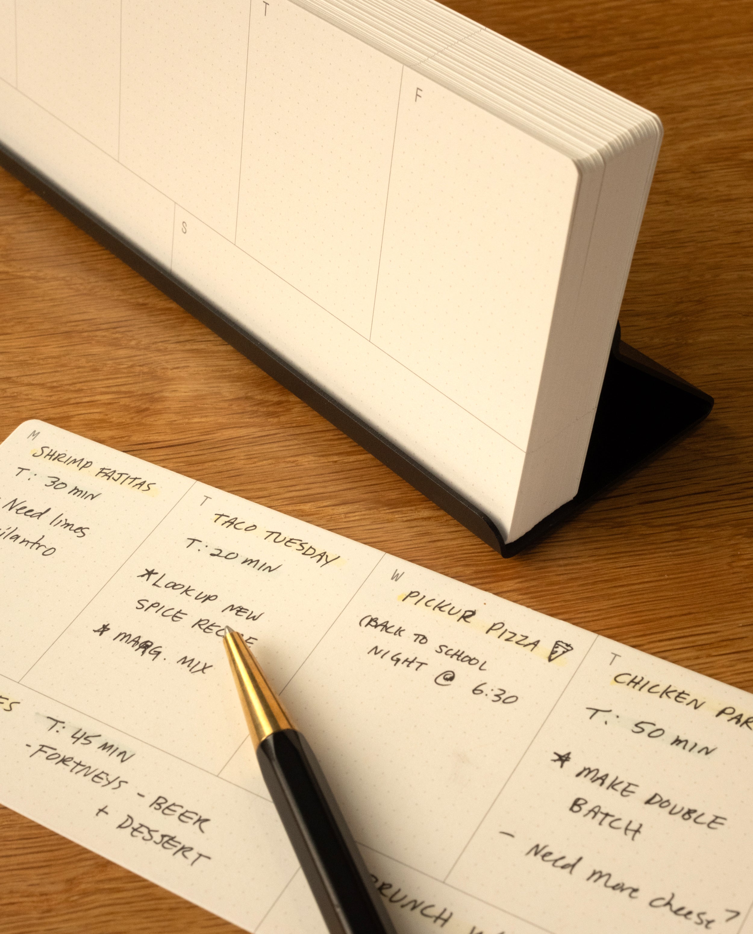 A gold and black pen rests on an open planner with handwritten meal plans—like taco Tuesday and pizza night—on a wooden desk next to the Analog Weekly Kit (Black Steel) weekly planner cards. A blank standing calendar is in the background.