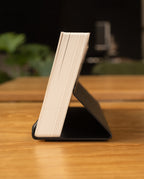 A book stands open on a wooden table, its pages fanned. In the background, a green plant and an Analog Weekly Kit (Black Steel) card holder are visible.