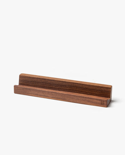The Analog Weekly Card Holder (Walnut) is a handcrafted, rectangular walnut stand made in Pennsylvania. Its groove holds items like Analog Weekly Cards, phones, or tablets. The visible wood grain and smooth finish add an elegant touch.