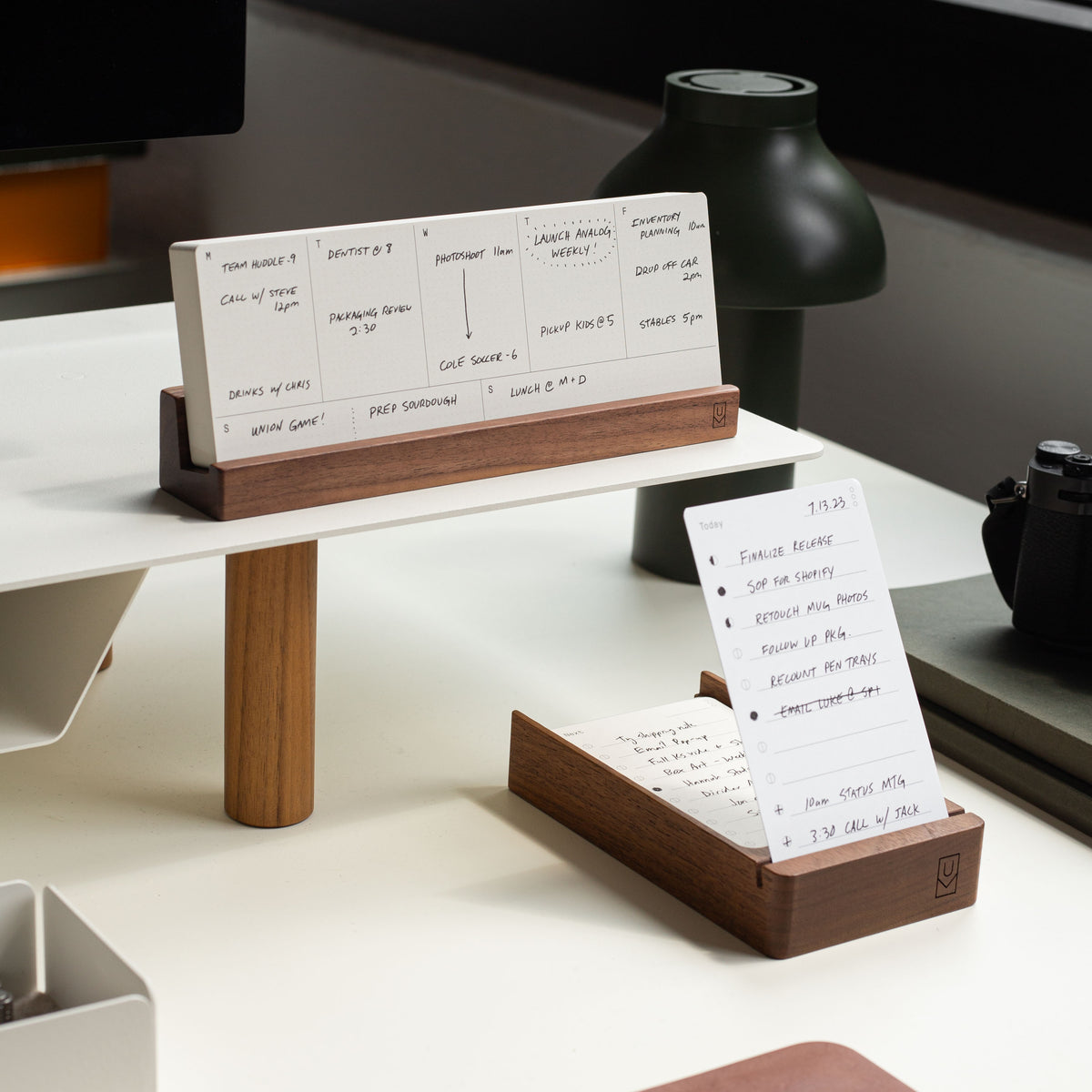 A workspace with wooden stands holding index cards with handwritten notes, set on a white desk shelf near a window. A lamp, camera, and monitor are also partially visible on the desk.