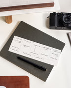 A black pen rests on a closed dark notebook, topped with a handwritten daily schedule. Nearby are a black camera, a white monitor riser, and part of a white desk. A walnut card holder elegantly displays neatly arranged weekly planner cards from the Analog Weekly Planning Kit (Walnut), adding an organized and refined touch to the workspace.