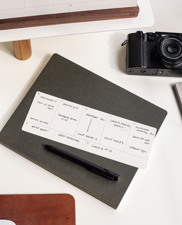 A black pen rests on a closed gray notebook with a handwritten card from the Analog Annual Refill (12 Task Packs + Weekly Cards) on top, while a camera and desk accessories complete the organized workspace look.