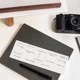 An Analog Weekly Card sits atop a closed dark notebook with a black pen, offering organized planning. In the background, a camera and part of a computer monitor are visible on the white desk.