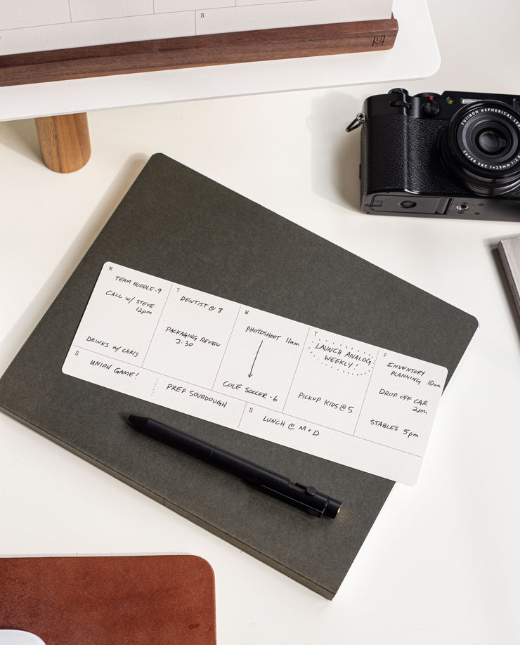 An Analog Weekly Card sits atop a closed dark notebook with a black pen, offering organized planning. In the background, a camera and part of a computer monitor are visible on the white desk.