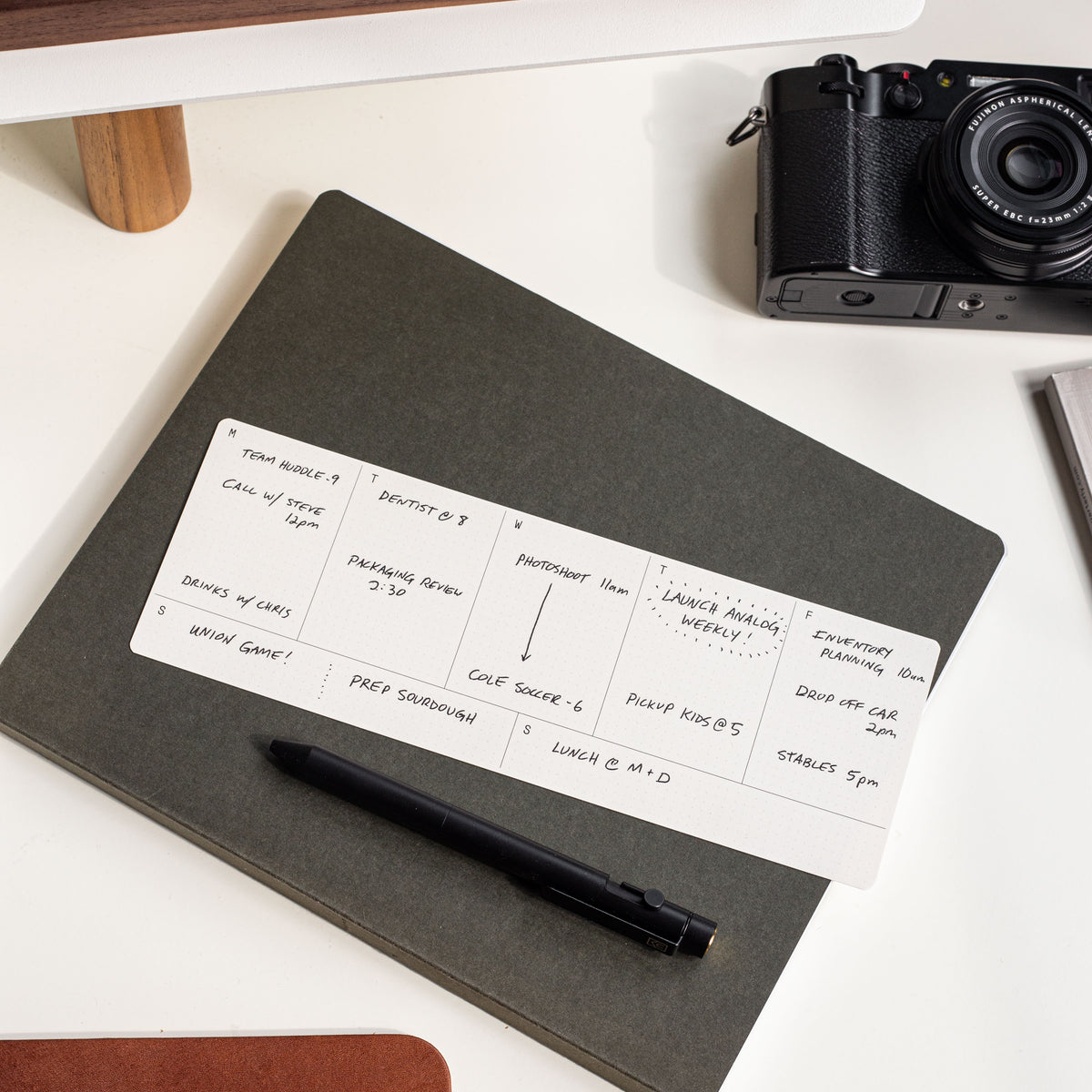 A black pen rests on a closed gray notebook, with a handwritten daily schedule card on top. Nearby are a camera and part of a calendar on a white stand. The scene is well-lit and organized.