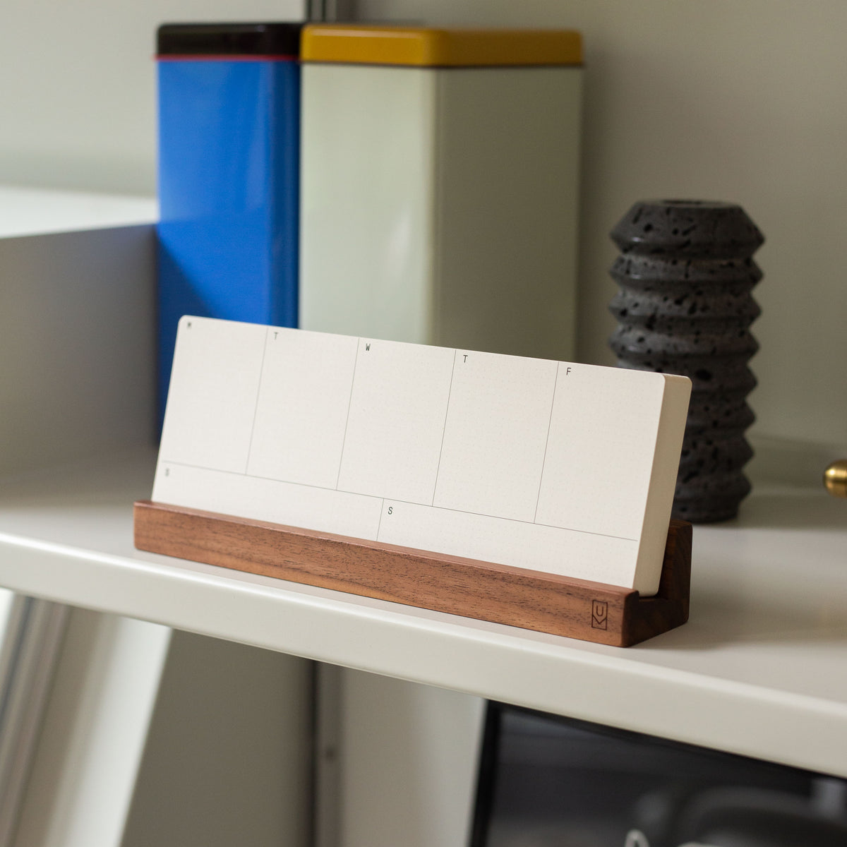 A blank, white, rectangular desk calendar sits on a wooden stand atop a white shelf, with colorful tins and a dark, textured object in the background.
