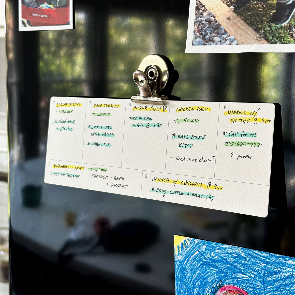 A handwritten weekly meal plan is clipped to a black fridge, surrounded by colorful family photos and childrens artwork. The plan lists daily dinners and events in different colored pens. Sunlight reflects off the fridge.