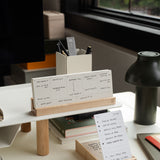 A modern desk displays the Analog Premium Weekly Kit (Maple) with a wooden organizer for handwritten note cards, a box of extra notes, pens in a holder, a green lamp, and natural light from a nearby window.