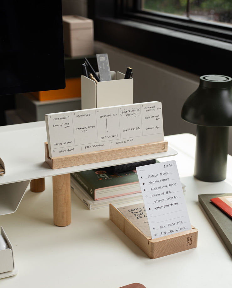 A modern desk displays the Analog Premium Weekly Kit (Maple) with a wooden organizer for handwritten note cards, a box of extra notes, pens in a holder, a green lamp, and natural light from a nearby window.