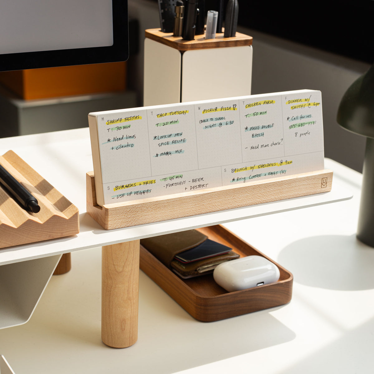 A modern desk setup features the Analog Weekly Planning Kit (Maple) displaying handwritten notes, with a tray for a phone and AirPods, and a pen holder. Sunlight streams in, creating a bright and organized workspace.
