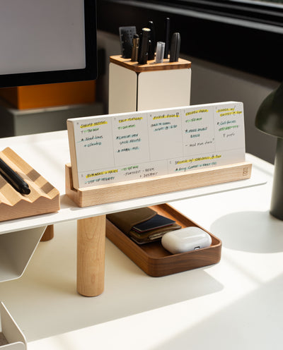 A modern desk setup features the Analog Weekly Planning Kit (Maple) displaying handwritten notes, with a tray for a phone and AirPods, and a pen holder. Sunlight streams in, creating a bright and organized workspace.