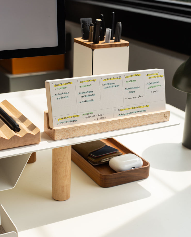 A modern desk setup features the Analog Weekly Planning Kit (Maple) displaying handwritten notes, with a tray for a phone and AirPods, and a pen holder. Sunlight streams in, creating a bright and organized workspace.