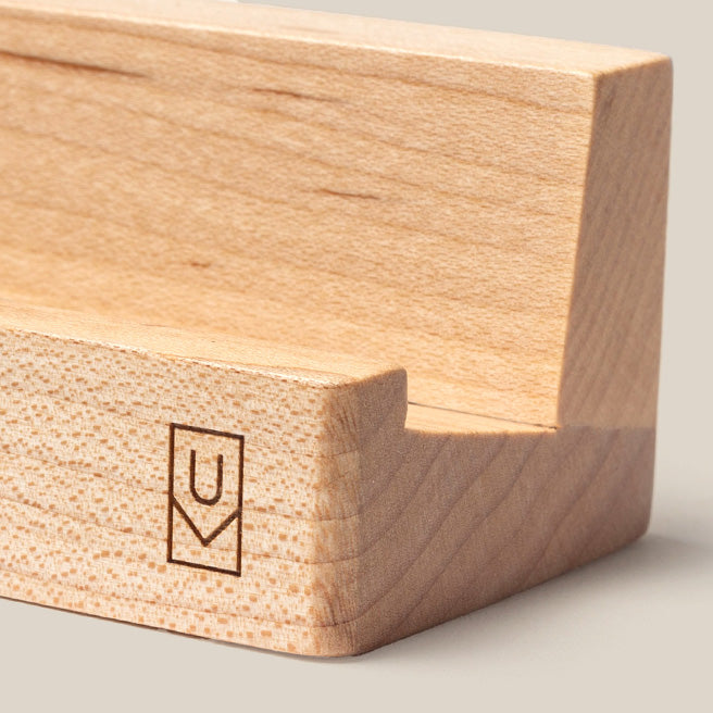 A close-up of a light wooden phone or tablet stand with a simple engraved logo featuring the letters VC inside a rectangle on the front corner.