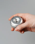 A hand holding the HMM Paperweight Pen Stand (Raw), a small, round, metallic aluminum object featuring concentric circular grooves and a central hole, against a plain gray background.