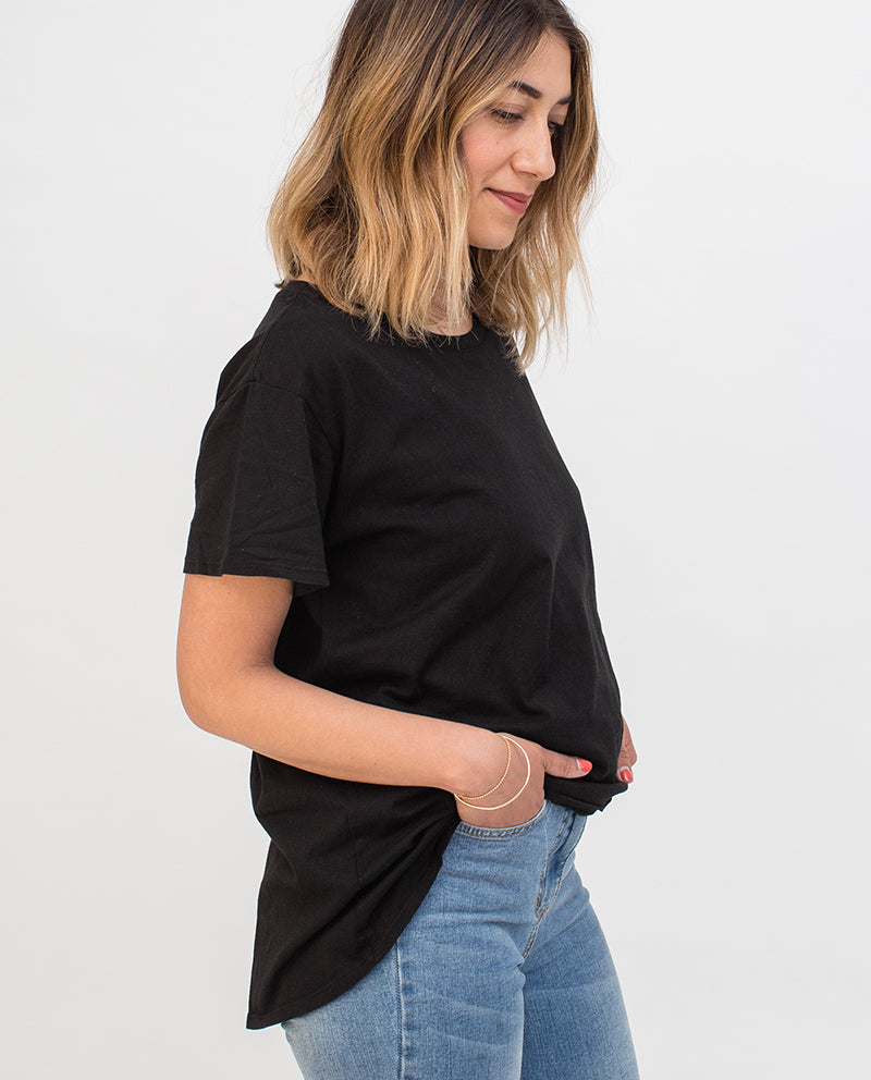 A woman with shoulder-length wavy hair stands in profile, wearing a black t-shirt and blue jeans. She has her hands in her pockets and is smiling slightly against a plain white background.